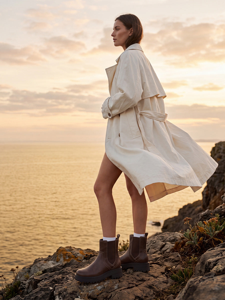 Model wearing brown chunky Chelsea boots with white socks and a cream trench coat on a rocky coast at sunset