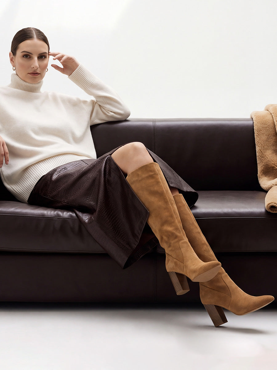 Model seated on a sofa wearing brown suede knee-high boots with a cream turtleneck and brown midi skirt