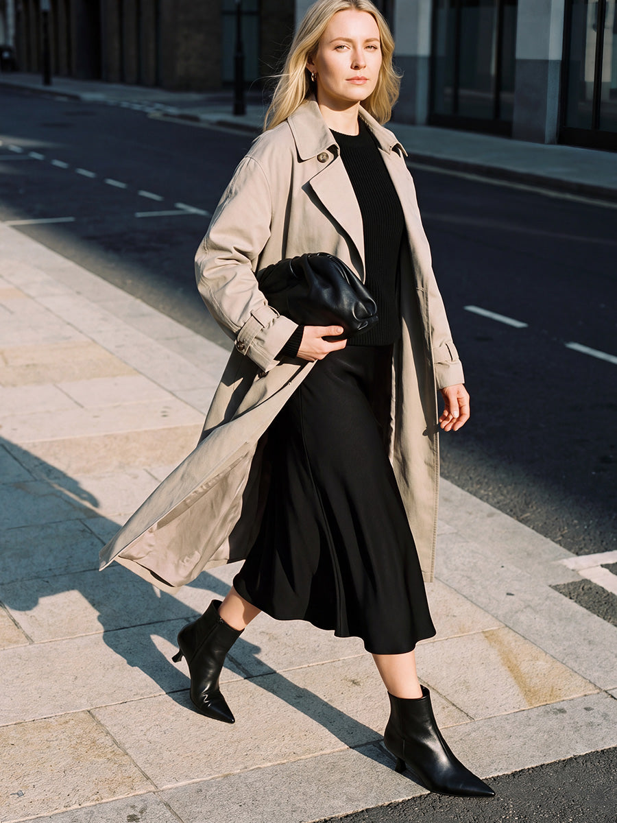 Model wearing black kitten-heel ankle boots with a black midi dress and beige trench coat while walking across a street