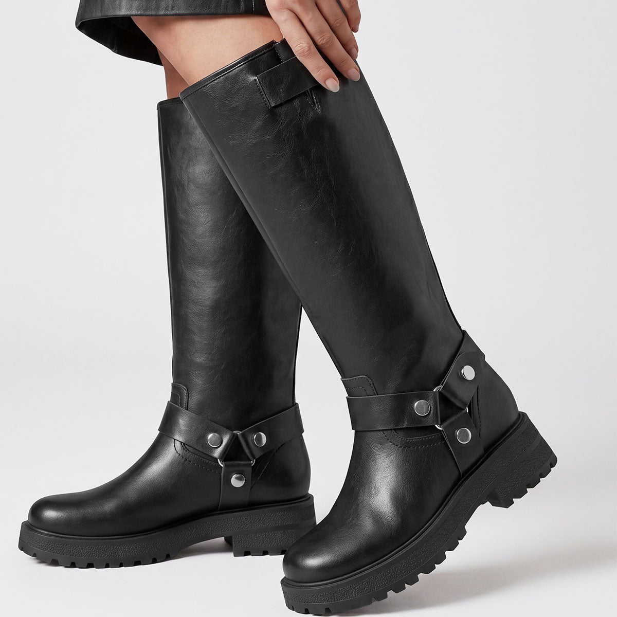 Black knee-high moto boots shown on foot, highlighting the tall shafts and ankle harness straps