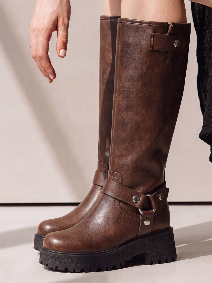 Close-up of brown knee-high moto boots showing the tall shafts, metal harness rings and chunky lug soles