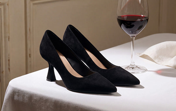 Black suede pointed-toe pumps displayed on a white tablecloth beside a wine glass