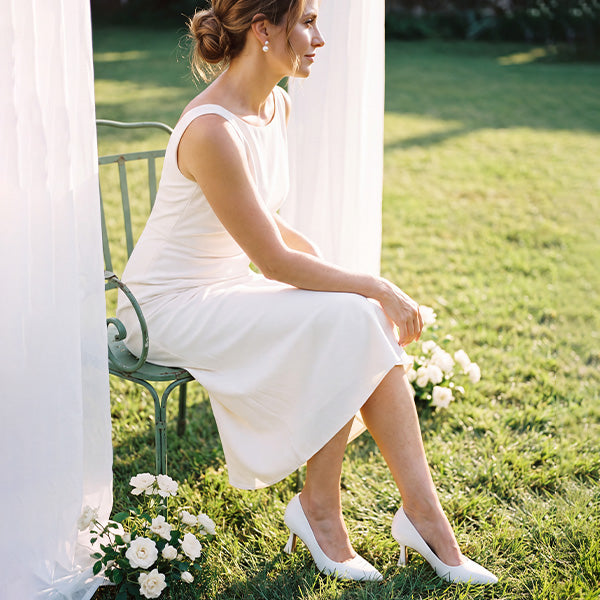 Model wearing white pointed-toe pumps with a white sleeveless dress while seated in a garden