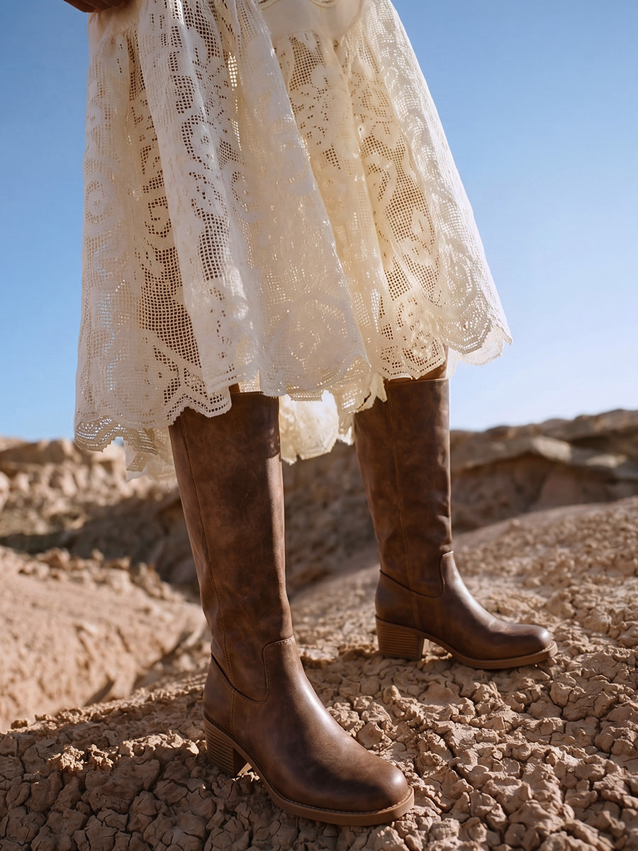 Brown square-toe knee-high boots styled with a cream lace dress on rocky ground